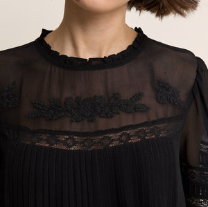 Close-up of a model wearing a black sheer mesh blouse with floral lace embroidery across the chest and a ruffled neckline.