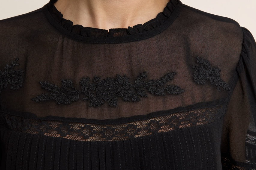 Close-up of a model wearing a black sheer mesh blouse with floral lace embroidery across the chest and a ruffled neckline.