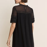 A woman wearing a black short-sleeve shift dress with sheer mesh sleeves and a lace-trimmed neckline, shown from the back against a plain beige studio background.