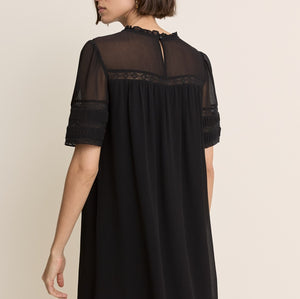 A woman wearing a black short-sleeve shift dress with sheer mesh sleeves and a lace-trimmed neckline, shown from the back against a plain beige studio background.