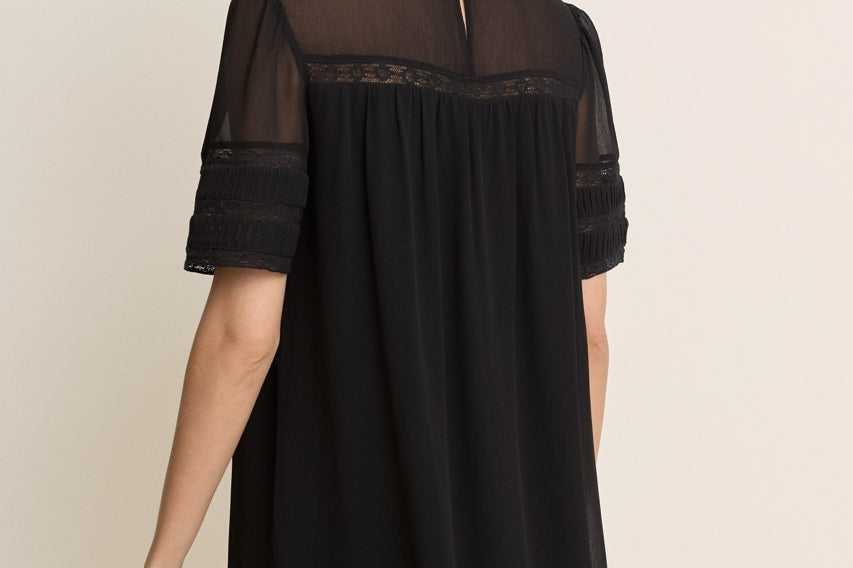 A woman wearing a black short-sleeve shift dress with sheer mesh sleeves and a lace-trimmed neckline, shown from the back against a plain beige studio background.