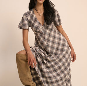 A woman models a short-sleeve plaid wrap dress with a V-neck, paired with tan knee-high boots, posed on a wooden block against a solid beige background.