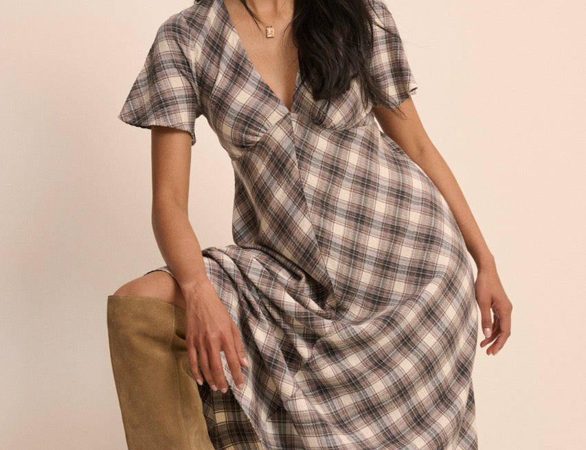 A woman models a short-sleeve plaid wrap dress with a V-neck, paired with tan knee-high boots, posed on a wooden block against a solid beige background.