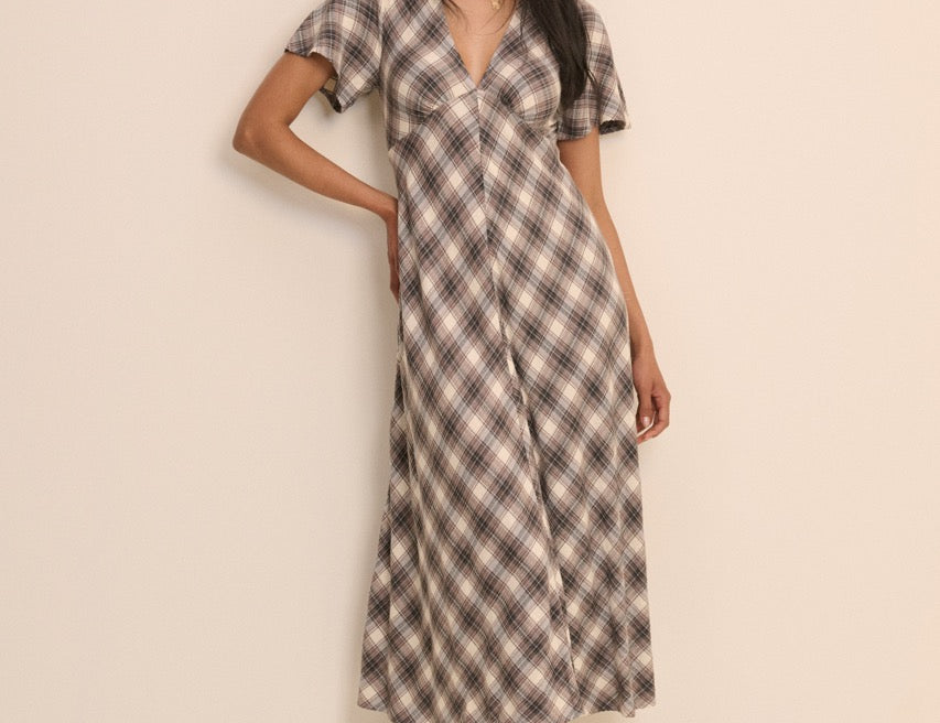 A woman modeling a short-sleeve midi dress with a brown, beige, and white plaid pattern against a plain beige studio background.