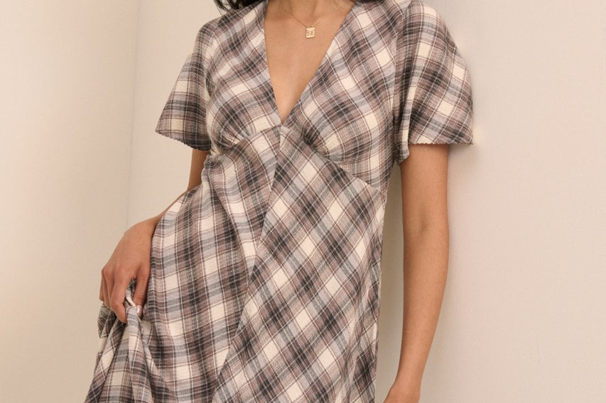 A woman modeling a short-sleeve plaid midi dress with a wrap-style V-neckline, photographed against a plain beige wall.