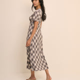 A woman modeling a short-sleeve midi dress with a brown and beige plaid pattern, standing against a solid light beige studio background.