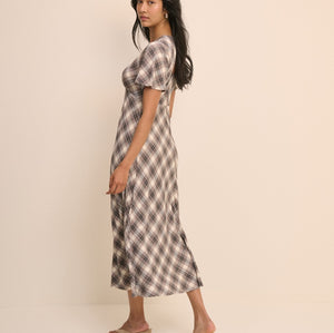 A woman modeling a short-sleeve midi dress with a brown and beige plaid pattern, standing against a solid light beige studio background.