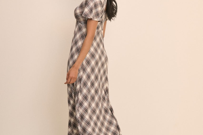 A woman modeling a short-sleeve midi dress with a brown and beige plaid pattern, standing against a solid light beige studio background.