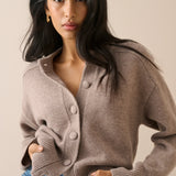 A woman wearing a taupe brown knit cardigan sweater with large buttons, paired with blue jeans, against a solid beige background.