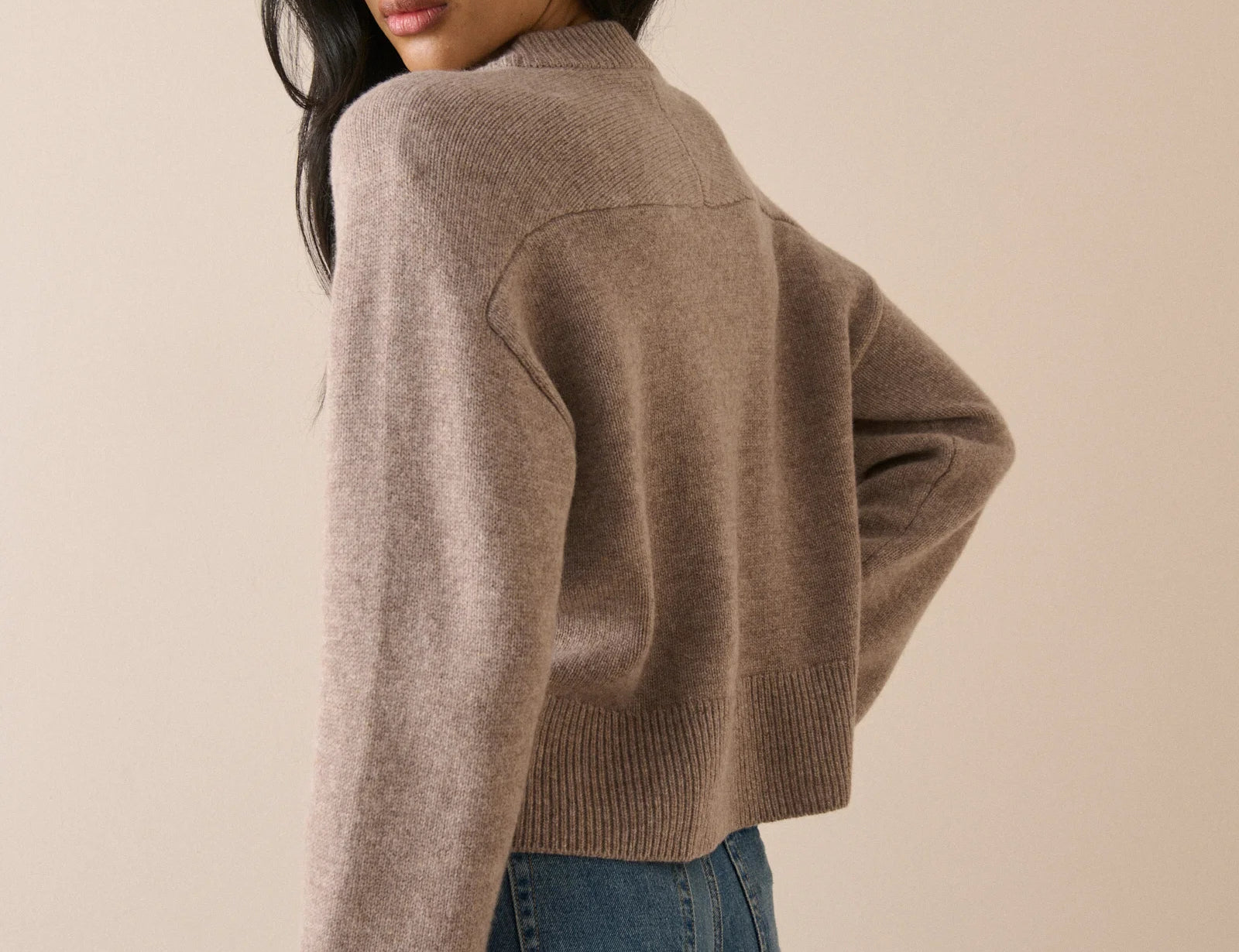 A woman wearing a cropped, long-sleeve knit sweater in a taupe-brown color, paired with blue jeans, photographed against a solid beige studio background.