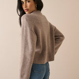 A woman wearing a cropped, long-sleeve knit sweater in a taupe-brown color, paired with blue jeans, photographed against a solid beige studio background.
