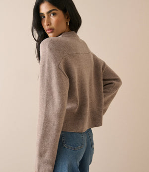 A woman wearing a cropped, long-sleeve knit sweater in a taupe-brown color, paired with blue jeans, photographed against a solid beige studio background.