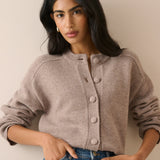 A woman wearing a taupe knit cardigan sweater with large buttons and blue jeans, posing against a neutral beige wall.