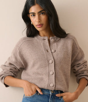 A woman wearing a taupe knit cardigan sweater with large buttons and blue jeans, posing against a neutral beige wall.