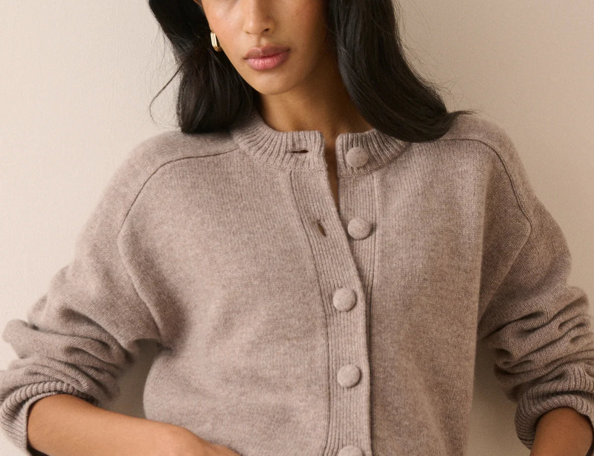 A woman wearing a taupe knit cardigan sweater with large buttons and blue jeans, posing against a neutral beige wall.