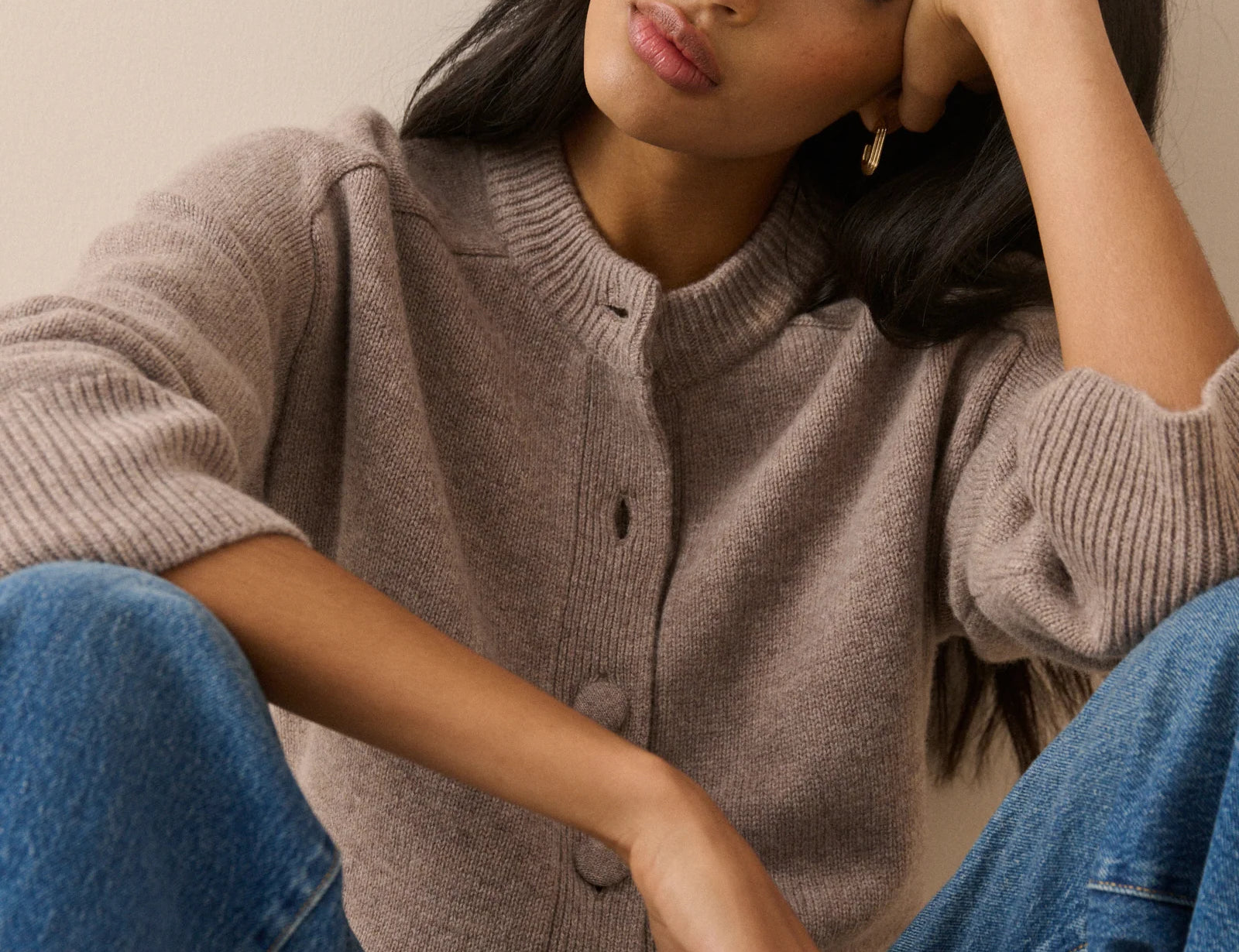A woman wearing a taupe ribbed knit cardigan sweater and blue jeans, posing with her head resting on her hand against a plain beige background.