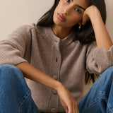 A woman wearing a taupe ribbed knit cardigan sweater and blue jeans, posing with her head resting on her hand against a plain beige background.