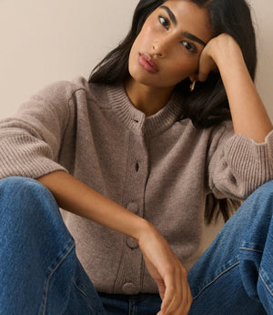 A woman wearing a taupe ribbed knit cardigan sweater and blue jeans, posing with her head resting on her hand against a plain beige background.