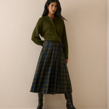 A woman stands against a plain beige studio wall wearing an olive green cardigan, a pleated green and navy tartan skirt, and black knee-high boots.
