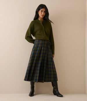 A woman stands against a plain beige studio wall wearing an olive green cardigan, a pleated green and navy tartan skirt, and black knee-high boots.
