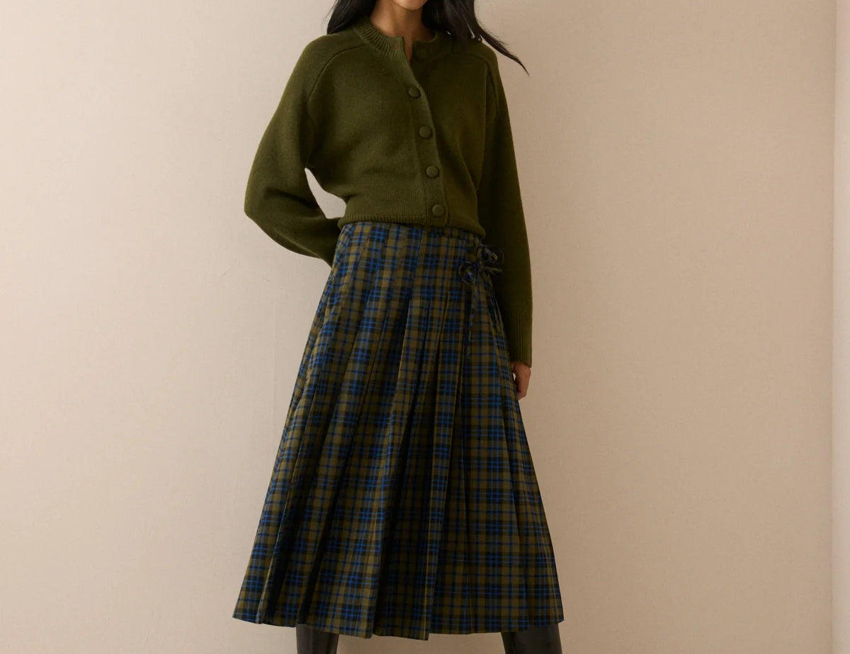 A woman stands against a plain beige studio wall wearing an olive green cardigan, a pleated green and navy tartan skirt, and black knee-high boots.