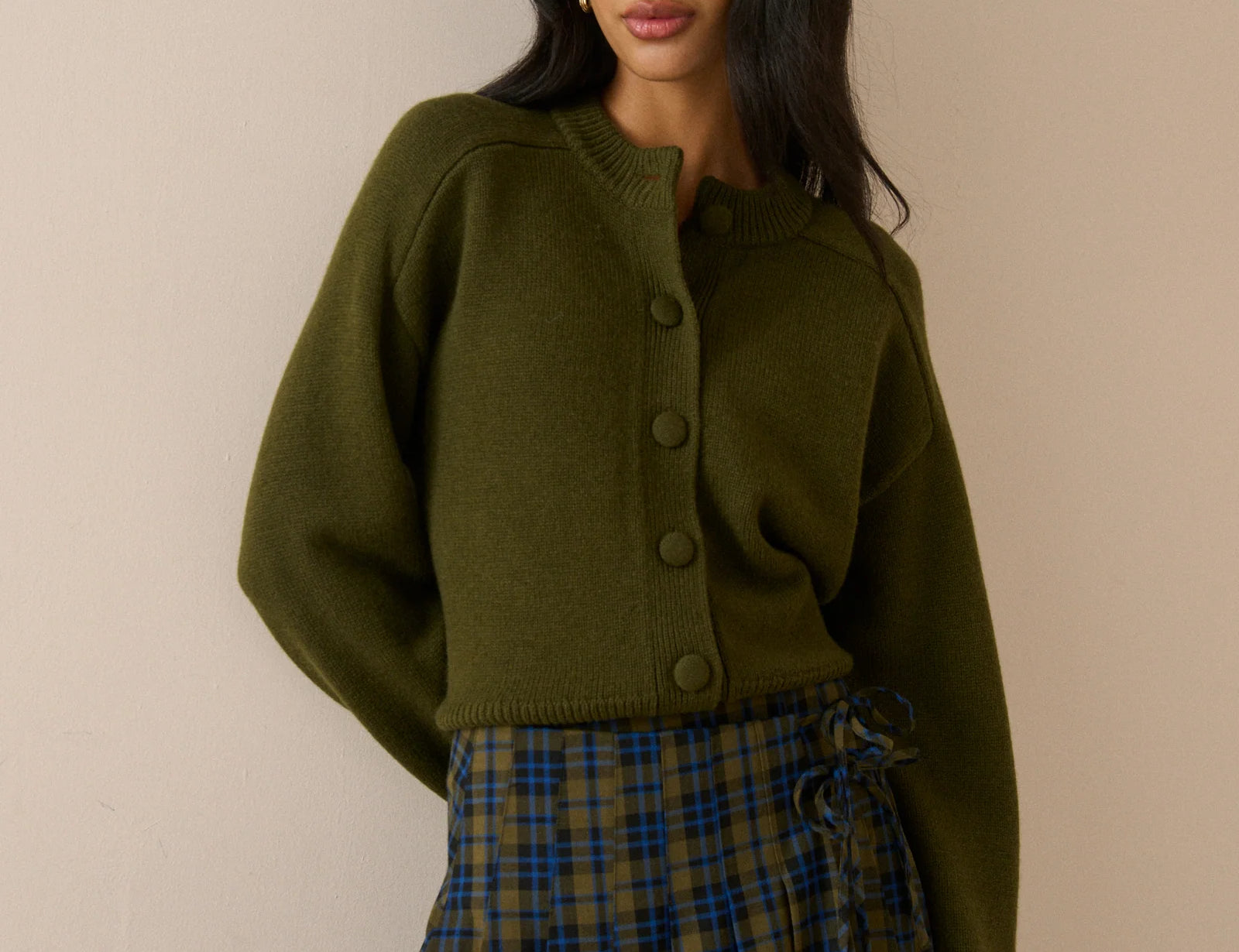 A woman modeling an olive green cropped button-front cardigan sweater paired with a blue and green plaid skirt, set against a plain beige wall.