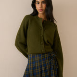 A woman modeling an olive green cropped button-front cardigan sweater paired with a blue and green plaid skirt, set against a plain beige wall.