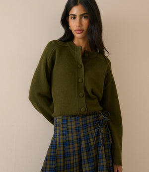 A woman modeling an olive green cropped button-front cardigan sweater paired with a blue and green plaid skirt, set against a plain beige wall.