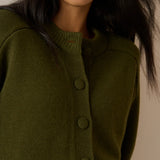Close-up of a woman wearing an olive green knit cardigan sweater with matching buttons, shown against a plain beige background.