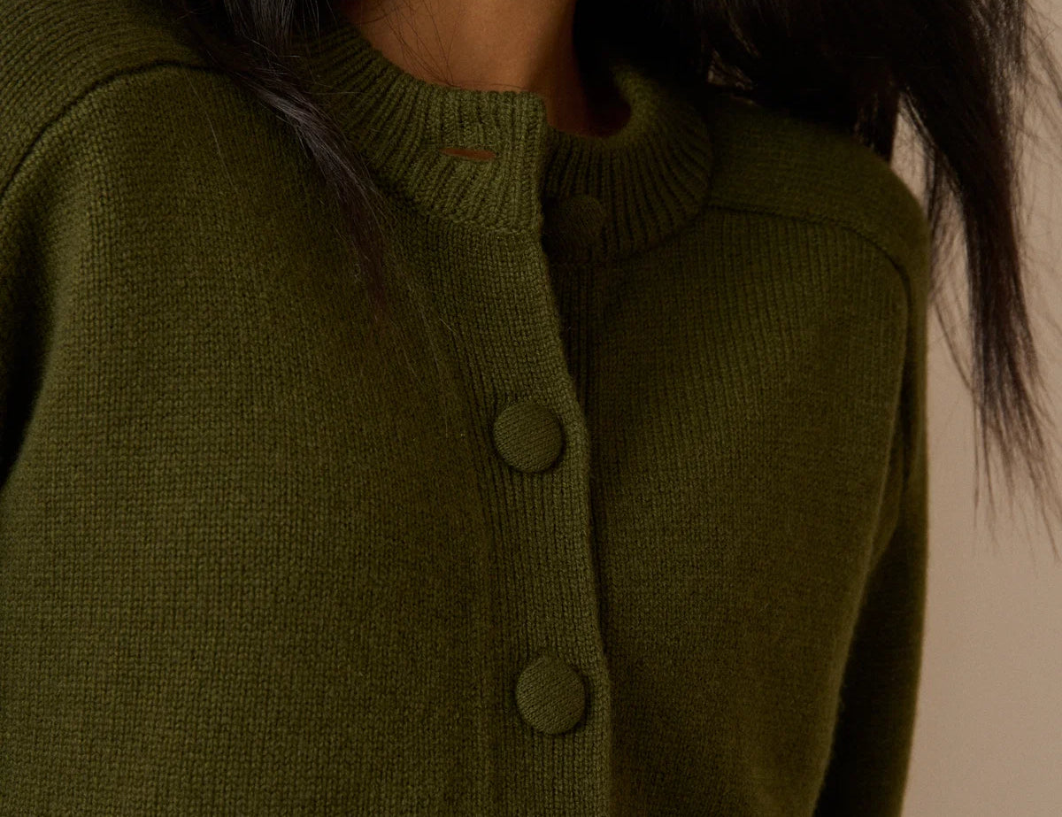 Close-up of a woman wearing an olive green knit cardigan sweater with matching buttons, shown against a plain beige background.