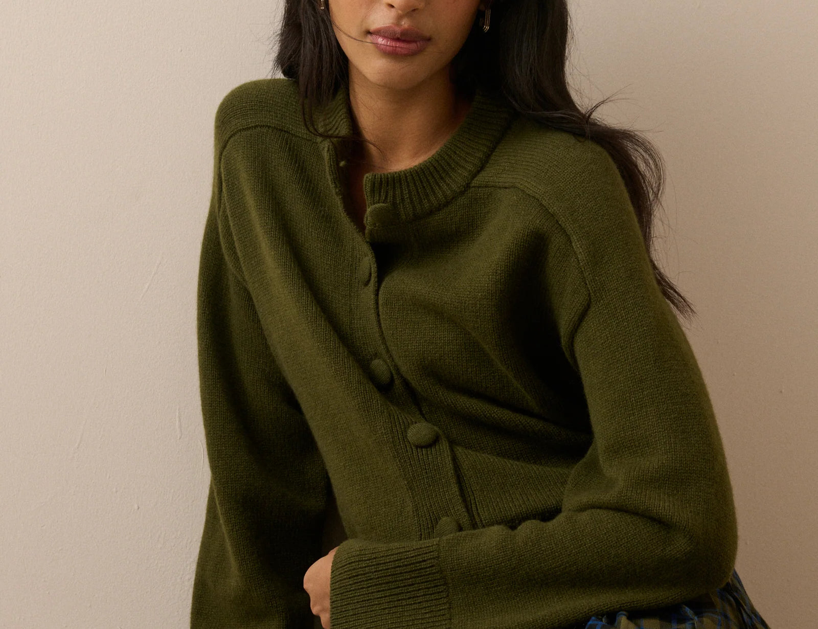 A woman wearing an olive green button-front cardigan sweater and a green plaid skirt, seated against a plain beige wall.