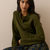 A woman wearing an olive green button-front cardigan sweater and a green plaid skirt, seated against a plain beige wall.