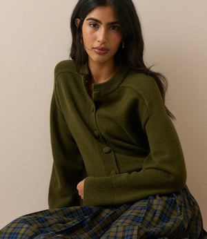 A woman wearing an olive green button-front cardigan sweater and a green plaid skirt, seated against a plain beige wall.