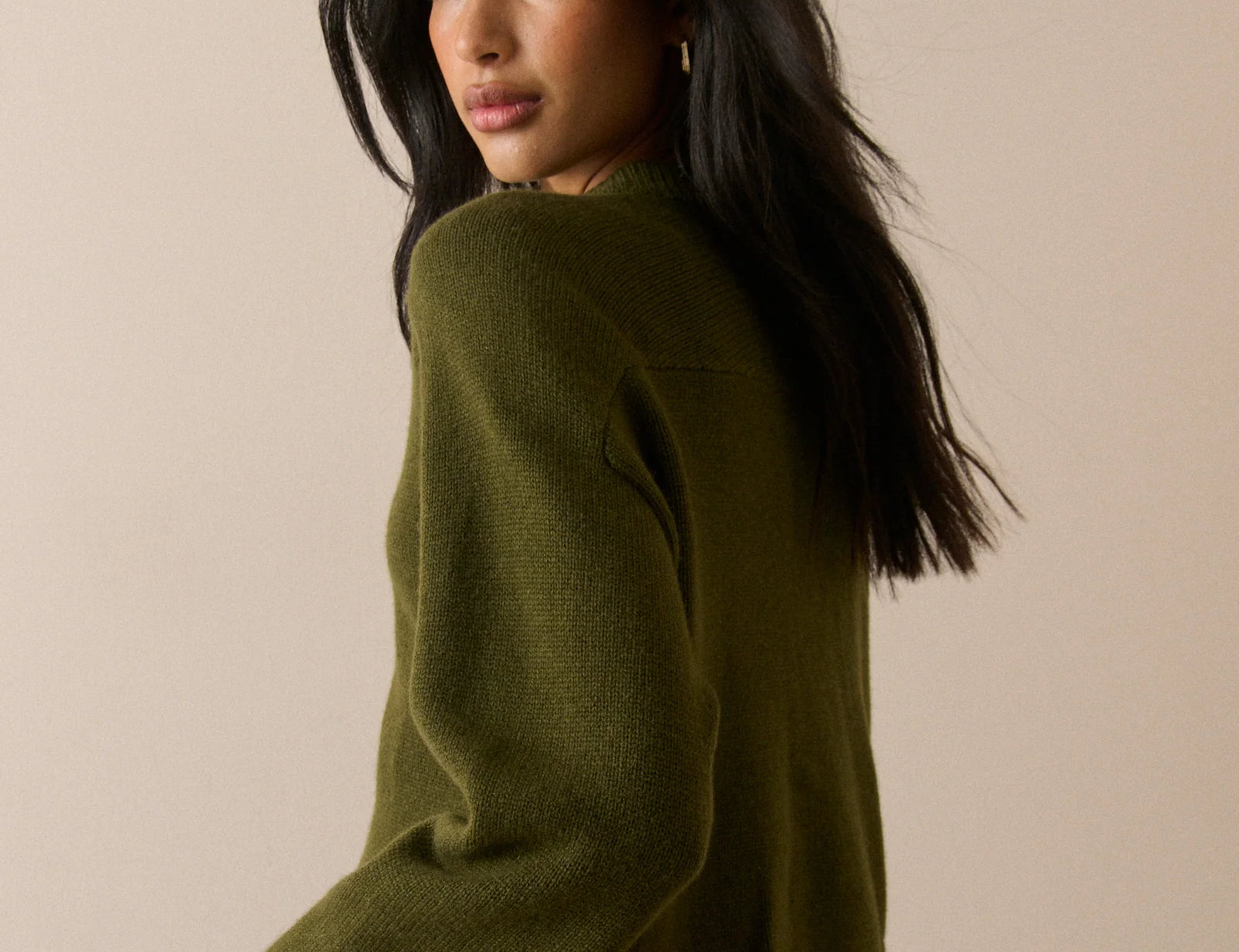 A woman with long dark hair is shown from a side-back angle wearing an olive green knit sweater and a blue plaid skirt against a plain beige studio background.