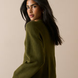 A woman with long dark hair is shown from a side-back angle wearing an olive green knit sweater and a blue plaid skirt against a plain beige studio background.