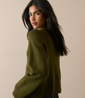 A woman with long dark hair is shown from a side-back angle wearing an olive green knit sweater and a blue plaid skirt against a plain beige studio background.