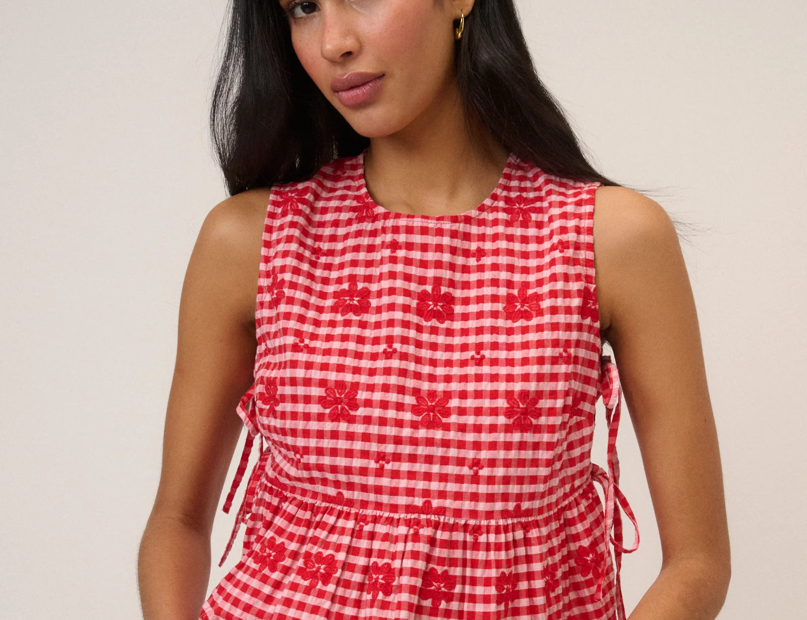 Woman wearing pink and red gingham top with flower detail and tie sides beige background