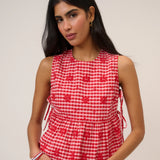 Woman wearing pink and red gingham top with flower detail and tie sides beige background