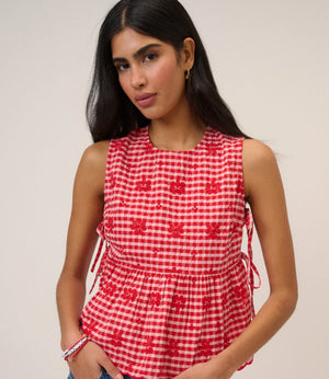 Woman wearing pink and red gingham top with flower detail and tie sides beige background