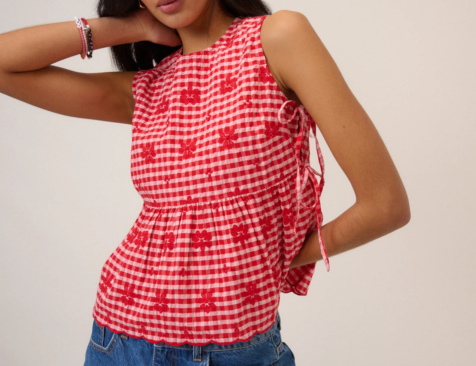 Woman wearing pink and red gingham top with flower detail and tie sides beige background