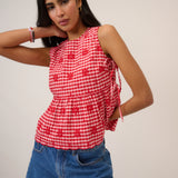 Woman wearing pink and red gingham top with flower detail and tie sides beige background