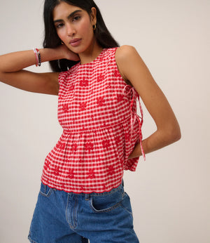 Woman wearing pink and red gingham top with flower detail and tie sides beige background