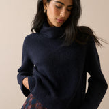 A woman wearing a dark navy blue knit turtleneck sweater, paired with a red and blue plaid skirt, against a solid beige background.
