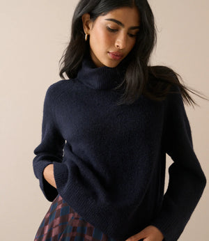 A woman wearing a dark navy blue knit turtleneck sweater, paired with a red and blue plaid skirt, against a solid beige background.