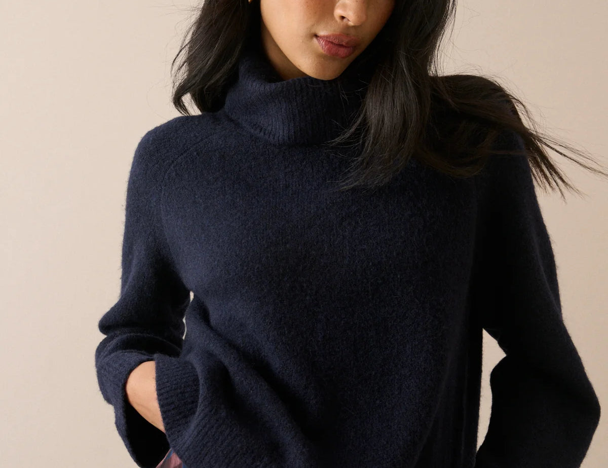A woman wearing a dark navy blue knit turtleneck sweater, paired with a red and blue plaid skirt, against a solid beige background.