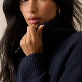 A close-up portrait of a woman wearing a dark navy blue ribbed knit turtleneck sweater, with her hand resting near her chin against a neutral beige background.