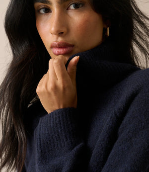 A close-up portrait of a woman wearing a dark navy blue ribbed knit turtleneck sweater, with her hand resting near her chin against a neutral beige background.