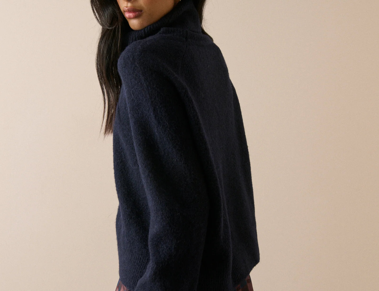 A woman wearing a dark navy blue fuzzy turtleneck sweater and a red and black plaid skirt, photographed against a solid beige background.