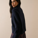 A woman wearing a dark navy blue fuzzy turtleneck sweater and a red and black plaid skirt, photographed against a solid beige background.