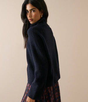 A woman wearing a dark navy blue fuzzy turtleneck sweater and a red and black plaid skirt, photographed against a solid beige background.